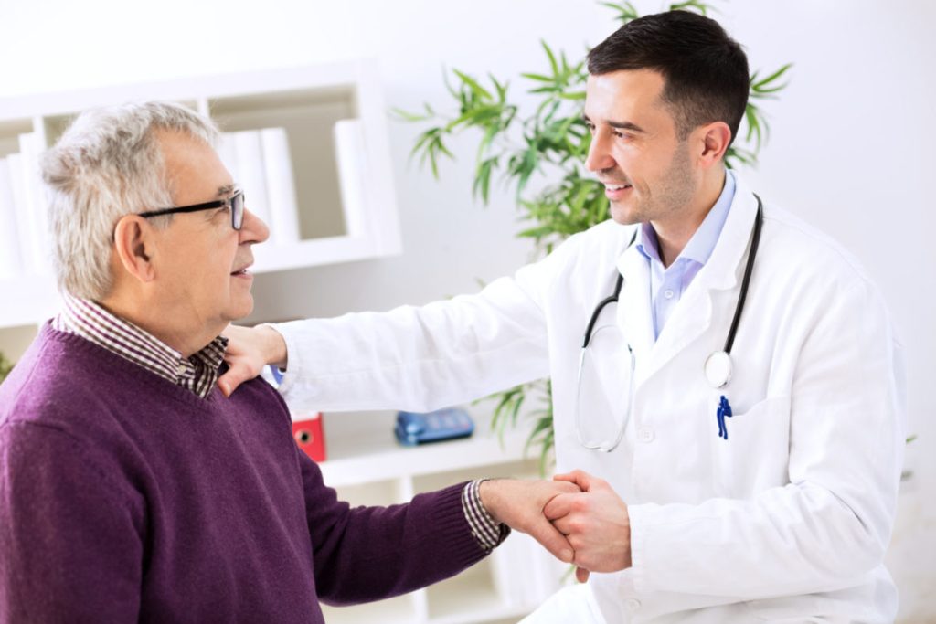Physician offering reassurance and support to an older male patient during a medical visit.