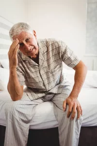 Older man sitting on edge of bed with his head in his hand, appearing fatigued or in discomfort, illustrating the physical and emotional side effects often associated with prostate cancer.